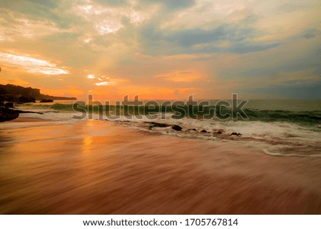 Seascape. Sunset time. Beach with rocks. Ocean with motion blur waves. Cloudy sky. Sunlight reflection on wet sand. Slow shutter speed. Soft focus. Tegal Wangi beach, Bali, Indonesia