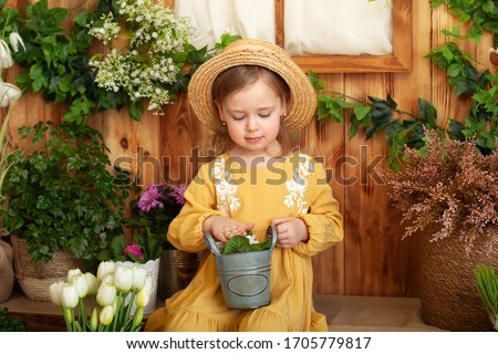 Little girl in dress and straw hat sits on porch of wooden house around green houseplants and flowers. Child planting spring flowers. Child taking care of plants. Little gardener plants plants in pot.