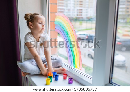 A little girl with blond hair is sitting in front of the window. The child is sad and looks out the window.