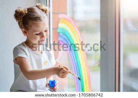 A little girl with blond hair draws a rainbow on the glass with paints. 