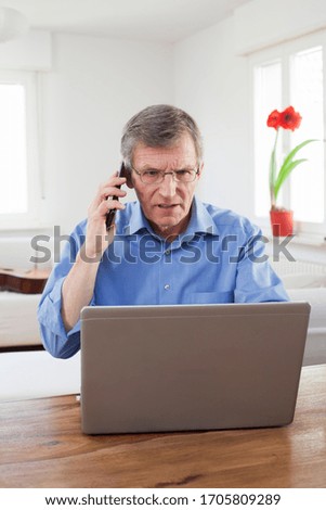 Serious mature businessman working at home with laptop while talking on cell phone - focus on the face