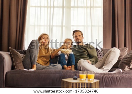 Front view of captivated family of mother, father and little daughter watching movie and eating popcorn sitting on sofa at home