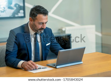The boss, who checks and takes notes at the computer in his own workroom in the office, the handsome hardworking boss with the beard in the suit, the bank manager, the team leader