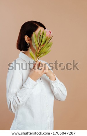 Portrait of young woman with black short hair holding large exotic flower, hiding her face. Girl wearing ring and earrings, hiding her face on beige background.
