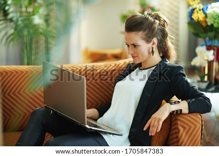 stylish 40 years old housewife in white blouse and black jacket in the modern living room in sunny day writing on a laptop while sitting on sofa.