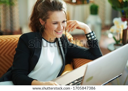smiling stylish middle age housewife in white blouse and black jacket in the modern living room in sunny day typing message on a laptop while sitting on sofa.