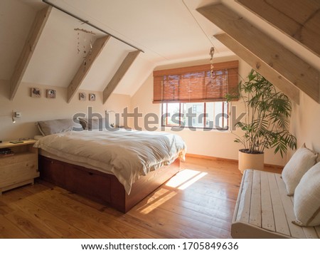 Cozy holiday bedroom for traveling guests, with beige bedding, white ceiling with wooden beams, green indoor plant and small bench with cushions Royalty-Free Stock Photo #1705849636