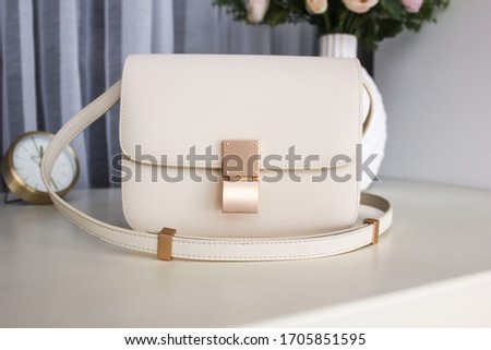 White women's leather bag with gold fittings on a white table with a vase, flowers and a clock in the background