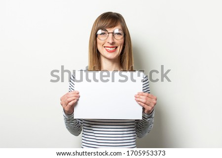 Portrait of a young friendly woman in a long-sleeve sweater and glasses with a smile, holding a blank sheet of paper on an isolated light background. Emotional face