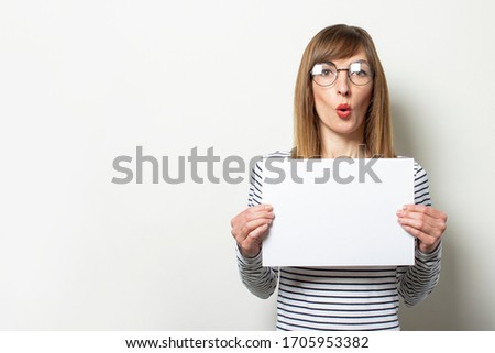 Portrait of a young friendly woman in a long-sleeve sweater and glasses with a surprised face holds a blank sheet of paper on light background. Emotional face. Gesture shock, surprise. Copy space