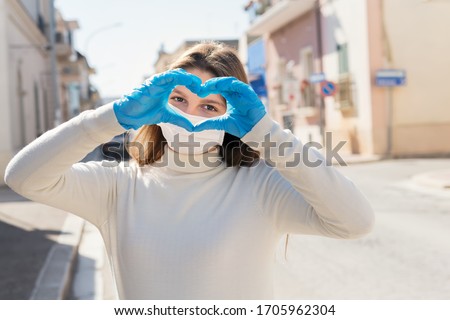 Girl wearing medical mask, and protective gloves, making heart shape sign by hands, is looking with hope, is middle of empty street. Valentines Day during pandemic of coronavirus