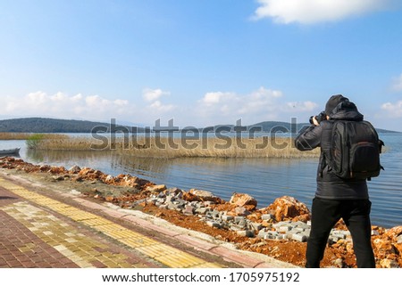 A young man with a photographer backpack is photographing outdoor near the seaside in a clear sky day. Photographing the wooden boat and the reeds inside the sea.