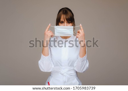 A girl doctor stands in a medical mask, isolated on a gray background.