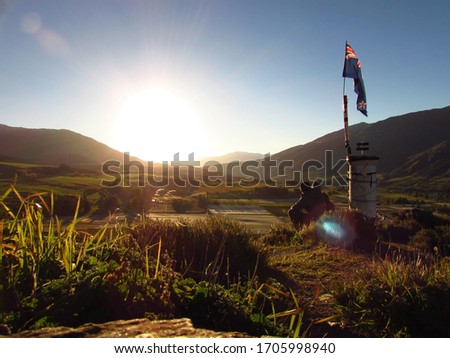 Couple sitting in sunrise on the hill under New Zealand flag. Mountains are surrounding the valley and vineyards are down below in the valley. The grass and hills are green and yellow.