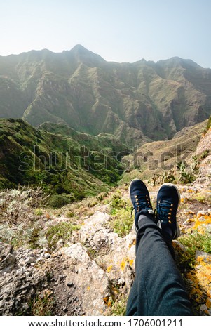 Legs of traveler sitting on high mountain cliff, enjoying scenery on mountain top. Pov view. Hiking freedom concept. Hiking in Anaga, legs of men in sneakers on a background of high mountains