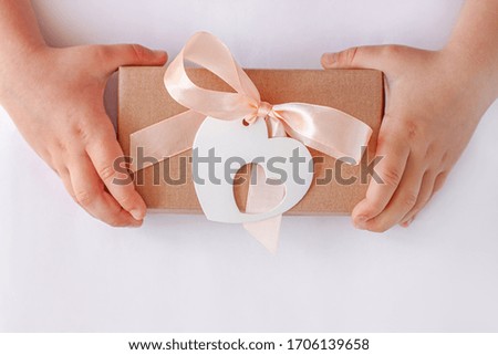Child's hands hold gift box with ribbon bow and heart card tag. Brown kraft paper packaging flat lay. Donation concept, Giving tuesday,love holidays Valentines Day.Minimalism creative style,copy space
