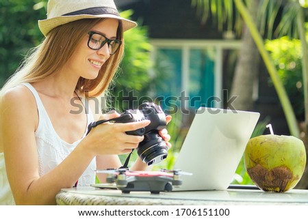 Female photographer inserting or removing a memory card in her professional dslr camera as she sits at a desk with her laptop at paradise beach green palms background