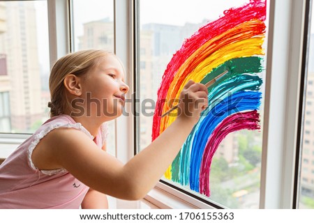 Kid painting rainbow during Covid-19 quarantine at home. Girl near window. Stay at home Social media campaign for coronavirus prevention, let's all be well, hope during coronavirus pandemic concept Royalty-Free Stock Photo #1706155363