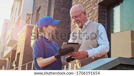 Old Caucasian male pensioner standing outdoors at his house and taking parcel from pretty young female postal worker courier. Woman delivering box to pensioner. Man signing. Mailing service.