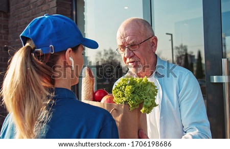 Rear of Caucasian young woman from delivery service in hat bringing packet with food to old man in glasses and giving it outdoors. Back view on female courier giving grocery deliver to grandfather. Royalty-Free Stock Photo #1706198686