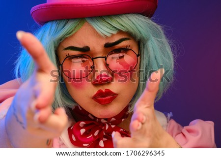Close-up portrait of a clown girl with makeup, blue hair, in a pink hat, pink dress and glasses. A clown shows different human emotions. April Fools Day concept. Circus performance with a clown.