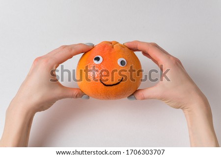 Grapefruit with funny face in female hands on white background. Orange with Googly eyes and painted smile.