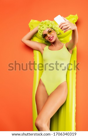 Top view of woman smiling, lying on inflatable mattress and taking selfie on orange