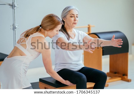 Older woman doing exercise on pilates chair equipment. Young female personal trainer helping senior woman. Workout in rehabilitation center, pilates studio. Seria photo with different angles.