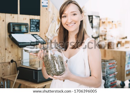Girl holds glass jar with hemp seeds. Woman with cotton reusable bag chooses and buys products in zero waste shop. Weighing dry goods in plastic free grocery store. Eco shopping at local business Royalty-Free Stock Photo #1706396005