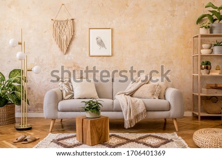 Stylish scandi compostion at living room interior with design gray sofa, wooden coffee table, shelf, cube, carpet, rattan pouf, plants, picture frame, table lamp and elegant accessories in home decor. Royalty-Free Stock Photo #1706401369