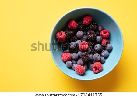 a beautiful light blue bowl of fruit with raspberries, blueberries and blackberries on yellow background, top view, studio shot