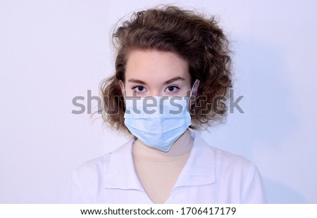 Studio portrait of curly girl in a medical lab coat wearing medical face mask. Protection against virus. Looking at the camera. Isolated on light gray background, close up. 
