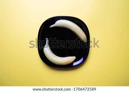 a modern pastel coloured image of two bananas an a decorative black square plate with pastel yellow background, flat lay, top view
