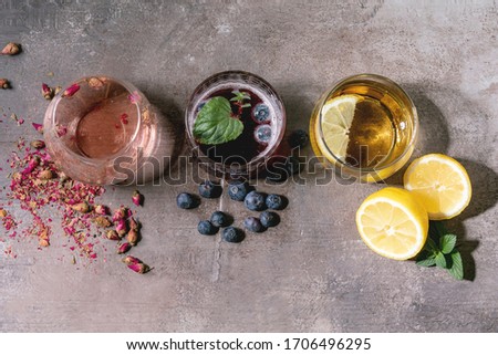 Different cocktail glasses with sparkling colorful drinks pink rose champagne, cider or lemonade with dry rose buds, lemon, blueberries and mint. Ingredients above. Texture background. Flat lay, space