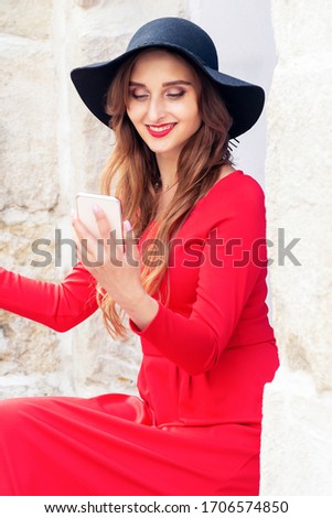 Woman is taking selfie wearing red dress and black hat at the city street.