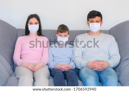 Mom, dad and son in protective medical masks sitting on couch at home and looking at camera, coronavirus pandemic quarantine. Worldwide global COVID-19 epidemic. Fighting with 2019-ncov infection.
