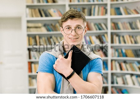 young smiling student in blue t-shirt and eyeglasses, holding black book in hands, and looking at camera while standing in front of big book shelves at modern reading hall in library