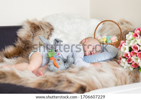  Little baby boy sleeping on a sofa on blue pillow and artificial fur, surrounded by easter decorations - bunny, basket with eggs and tulips