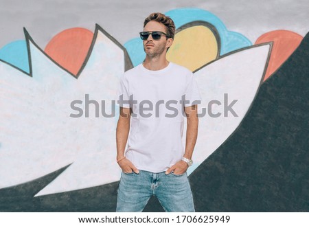  Young handsome guy is standing on a graffiti wall background. Man is wearing white, empty basic t-shirt without logo. Horizontal mock-up with light flare.