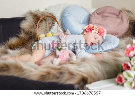 Little baby girl in flower crown, sleeping on a sofa on blue pillow and artificial fur, surrounded by easter decorations - bunny, tulips and basket with eggs