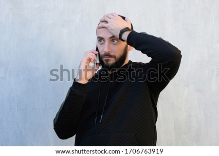 The guy is talking on the phone against a gray wall. The guy holds the phone near his ear and holds his head with other hand. Reporting bad news over the phone, surprise, tedious phone conversations