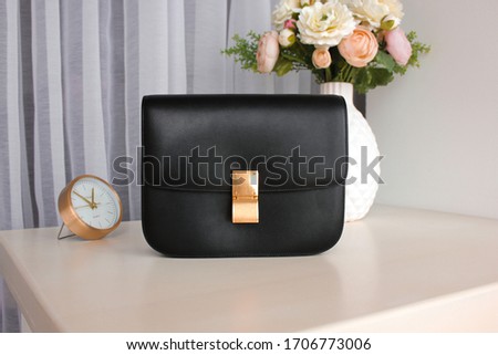 Black women's leather bag with on a white table with a vase, flowers and a clock in the background