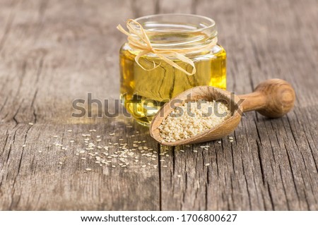 Glass jar of sesame oil and raw sesame seeds in wooden shovel with burlap sack on wooden table. Uncooked sesame background concept with copy space Royalty-Free Stock Photo #1706800627