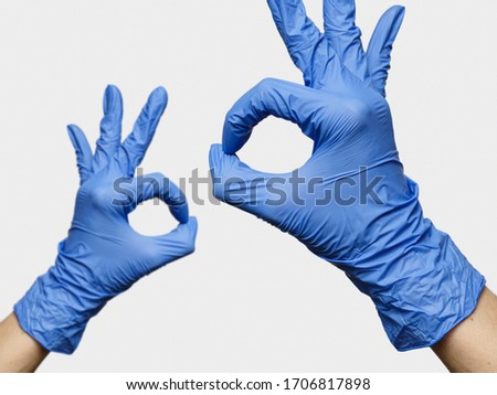 'OKAY' signs by female hands in blue medical gloves close and far away over light gray background. Closeup