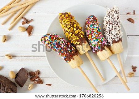 Frozen chocolate dipped banana pops with sprinkles, nuts and coconut. Overhead view table scene on a wood background. Royalty-Free Stock Photo #1706820256