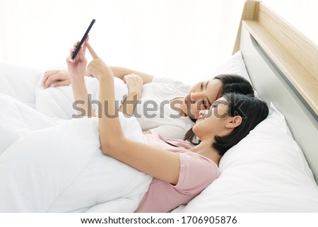 Asian young sisters lovely couple sitting on white bed and smiling together for Social online with smartphone in bedroom. Homosexual and asian Lesbian in relationship. ็Holiday and Technology Concept