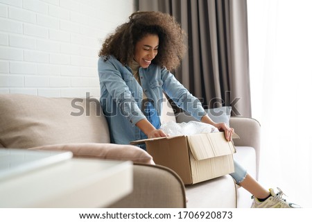 Excited african young woman shopper opening parcel box at home. Happy satisfied ethnic girl customer receiving online shop purchase by postal shipping, unpacking delivery receiving gift sit on sofa. Royalty-Free Stock Photo #1706920873