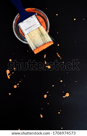 A brush and a glass jar full of paint lie on a dark surface, which is full of orange spots of paint - a picture with a shallow depth of field as a symbol of art and stability