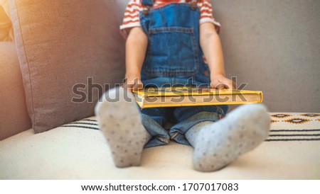 Little, cheerful, three years old boy on the sofa, with open book in front of him. Cute little boy is reading book. Shot of an adorable little boy reading a book while relaxing on the sofa at home