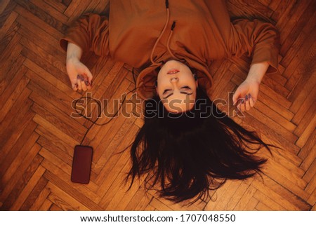 The girl lies on the floor and listens to music through headphones. Girl at home listening to music, top view. A woman, a brunette, lies on the floor and relaxes, sings.